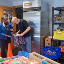 A woman hands a tote bag full of food to an older man in a room with shelves of fresh produce