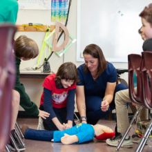 Nurse educator Sara Cohen kneels on the classroom floor with a child practicing the Heimleich maneuver on a doll.