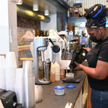 A woman wearing a black uniform shirt and blue and black braided hair pours coffee beans behind the counter of a coffee shop.